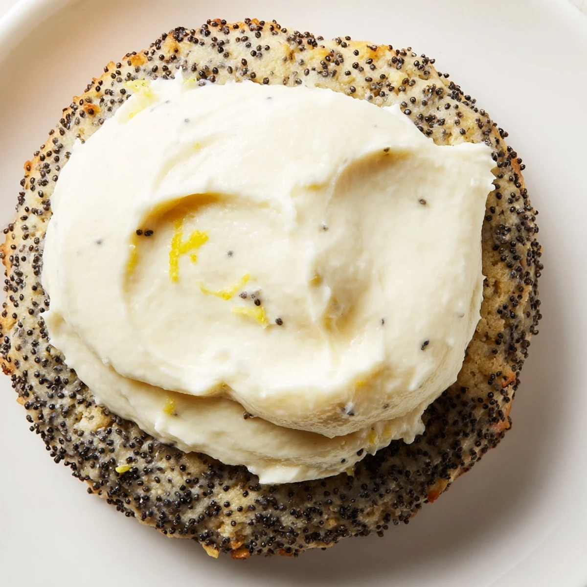 Golden lemon poppy seed cookies topped with fluffy white cheesecake frosting and sprinkled with poppy seeds on a white plate